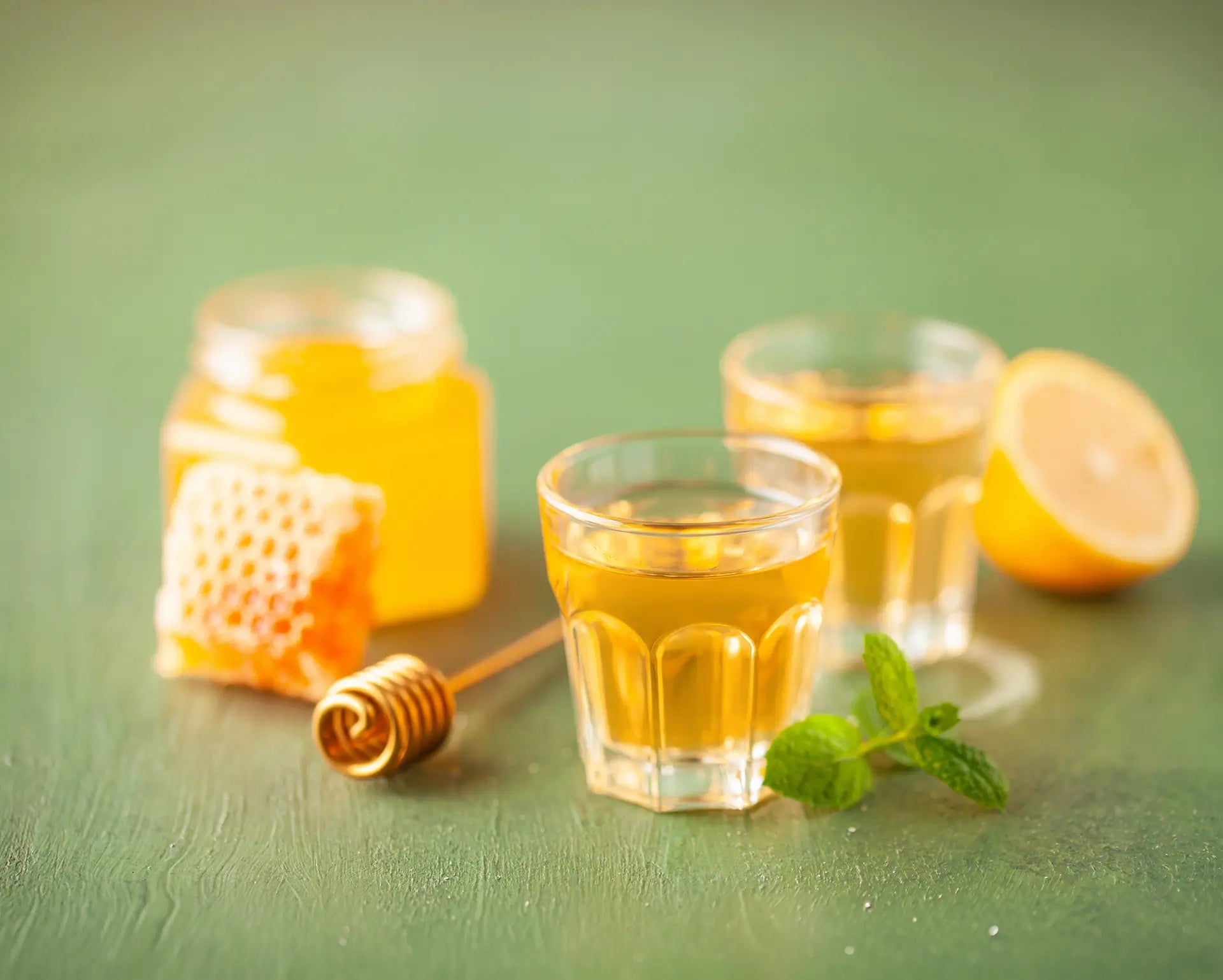 Frischer Bio-Honig im Glas und Wabenstück der Bioland-Imkerei Honigtreu, serviert mit zwei Gläsern Honiglikör, Zitrone und Minze auf grünem Holztisch.